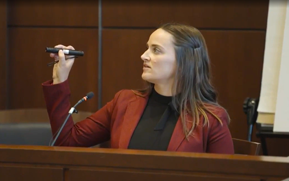 A woman in a courtroom setting raising a black object, possibly a recording device or microphone, while sitting behind a wooden barrier.