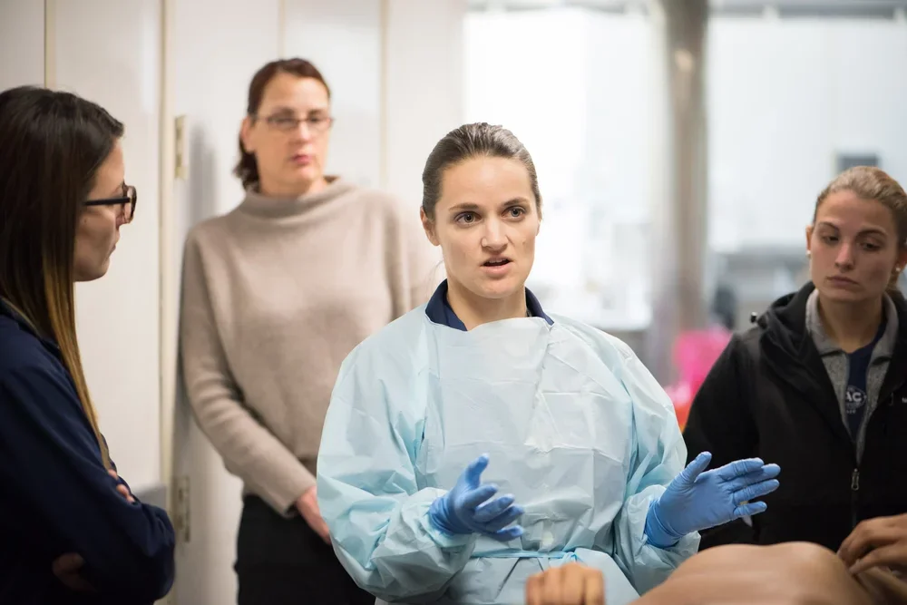 A female medical professional or instructor wearing a gown and gloves speaking to a group of women in a morgue setting.