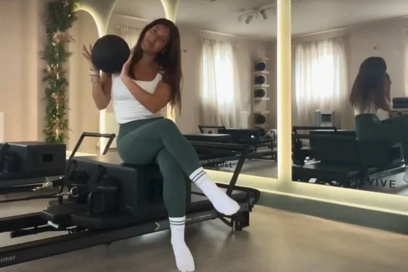 A woman sitting on a Pilates reformer machine, holding a black exercise ball, in a fitness studio with a large mirror reflecting her and the room.