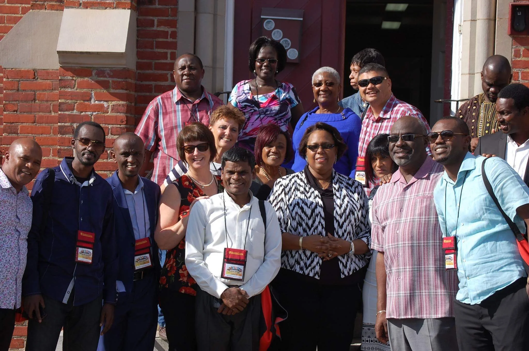 Group of men and women standing together outside a brick building, smiling for a group photo, some wearing sunglasses and ID badges.