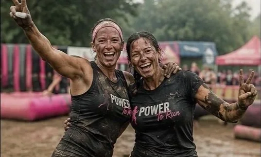 Two women smiling and celebrating with muddy faces after a race, wearing black shirts that say "POWER to Run," outdoors with pink inflatable barriers in the background.