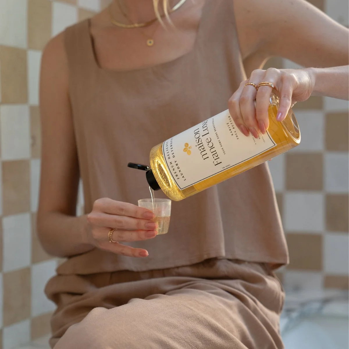 A woman pouring Faisence Luxe shea butter body oil from a clear bottle into a small shot glass.