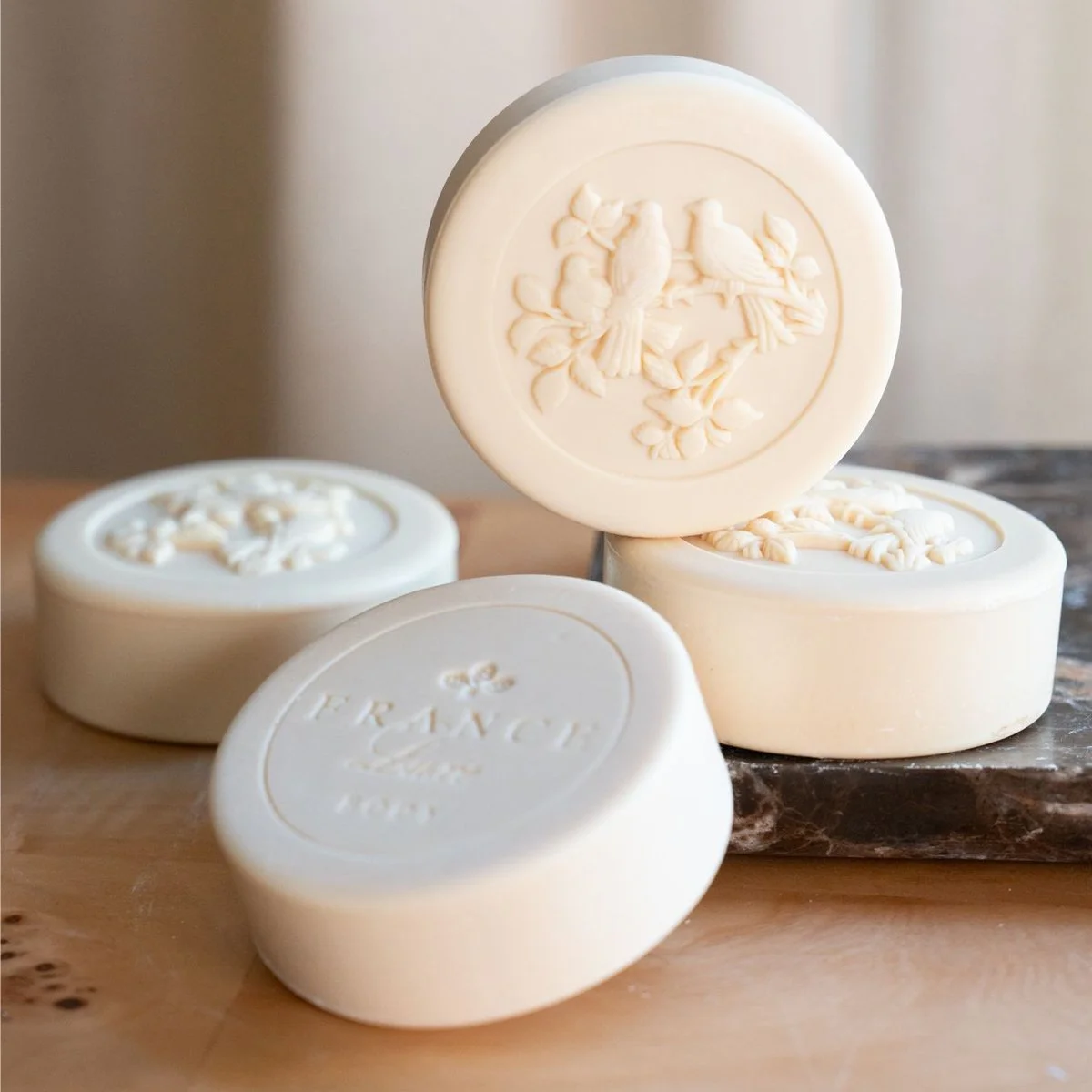 Four ivory-colored soap bars with embossed floral and bird designs, placed on a wooden and marble surface.