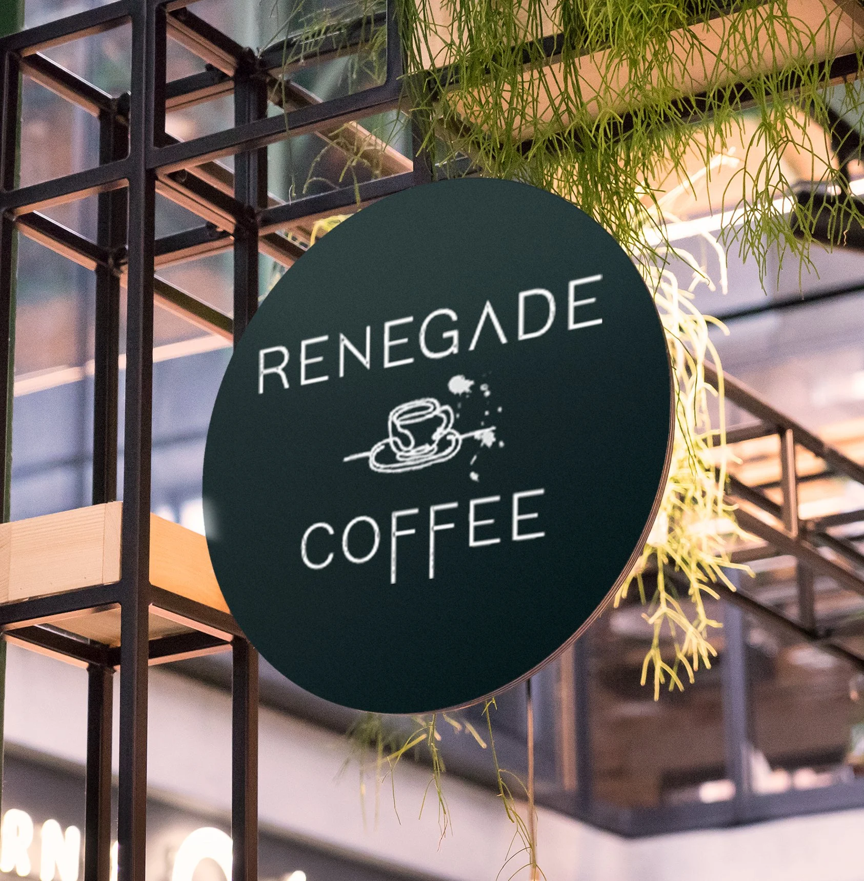 A black grid display with a round black sign that reads 'RENEGADE COFFEE' with a coffee cup illustration. Green plants hang above and around the sign indoors.