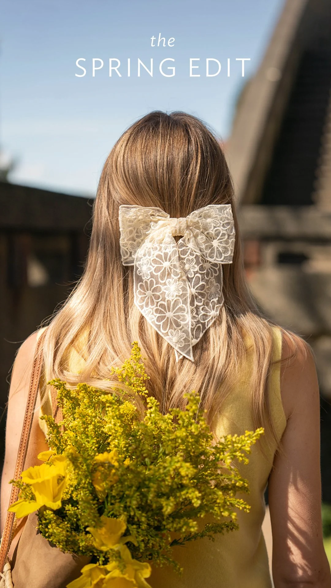 A woman with long blonde hair wearing a large lace bow in her hair, holding a bouquet of yellow flowers, standing outdoors on a sunny day.