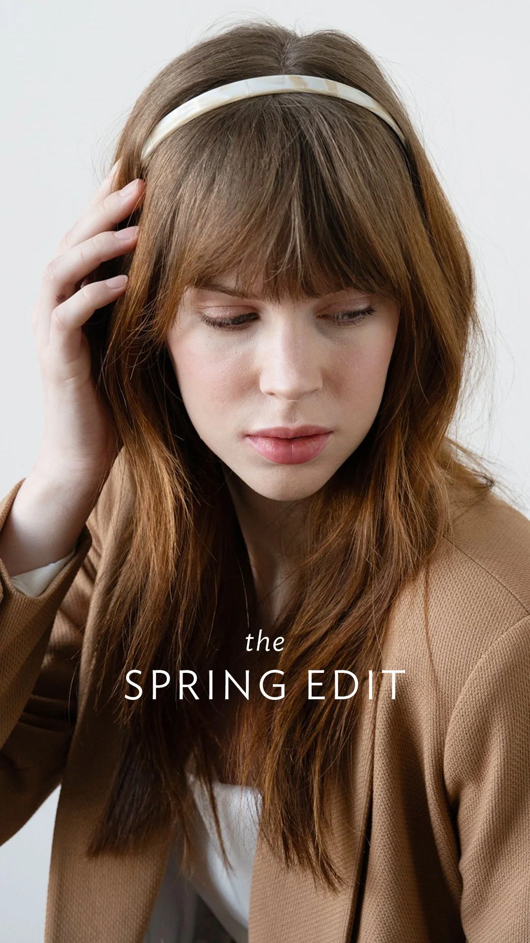 A woman with long red hair and bangs, wearing a beige blazer and white top, touches her head with her right hand, looking down gently. The text on the image reads "the SPRING EDIT."