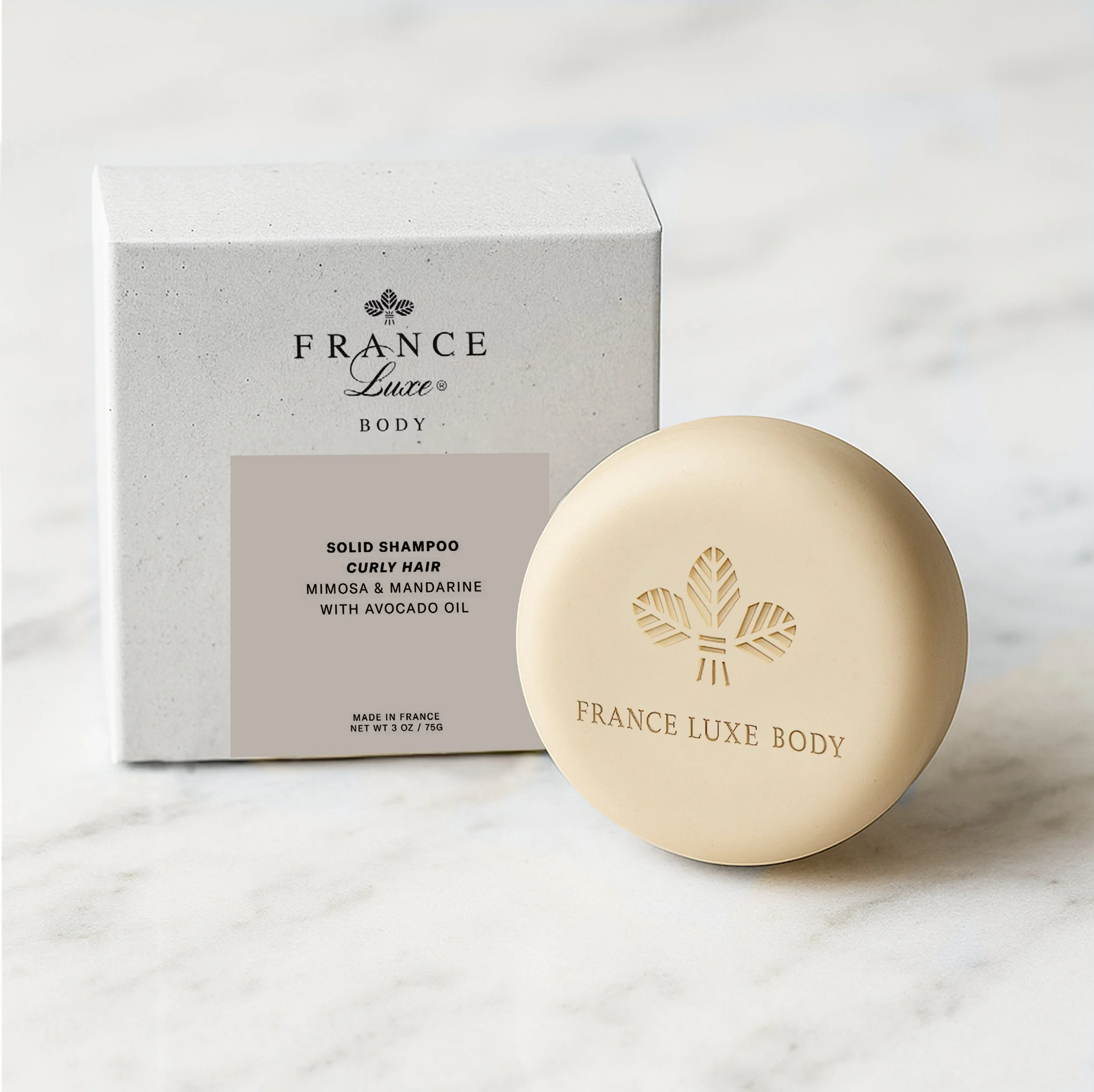 Close-up of a white box labeled 'France Luxe Body Solid Shampoo Curly Hair' and a round beige soap with a floral logo next to it. The background is white.