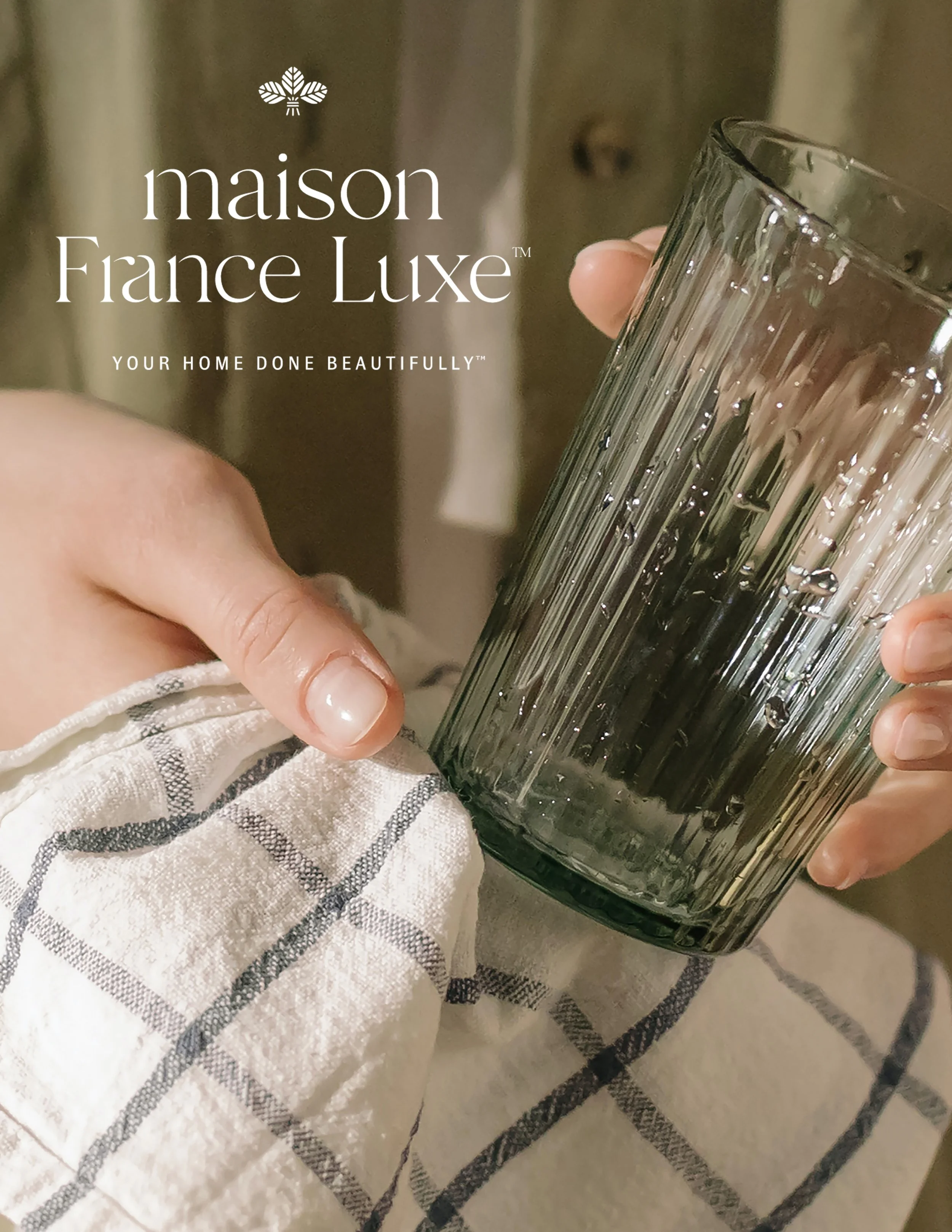 Close-up of a hand holding a tall, clear glass with a textured vertical line pattern. The background is blurred, showing a person in striped clothing. The text overlay reads: 'maison France Luxe - YOUR HOME DONE BEAUTIFULLY' with a small decorative f