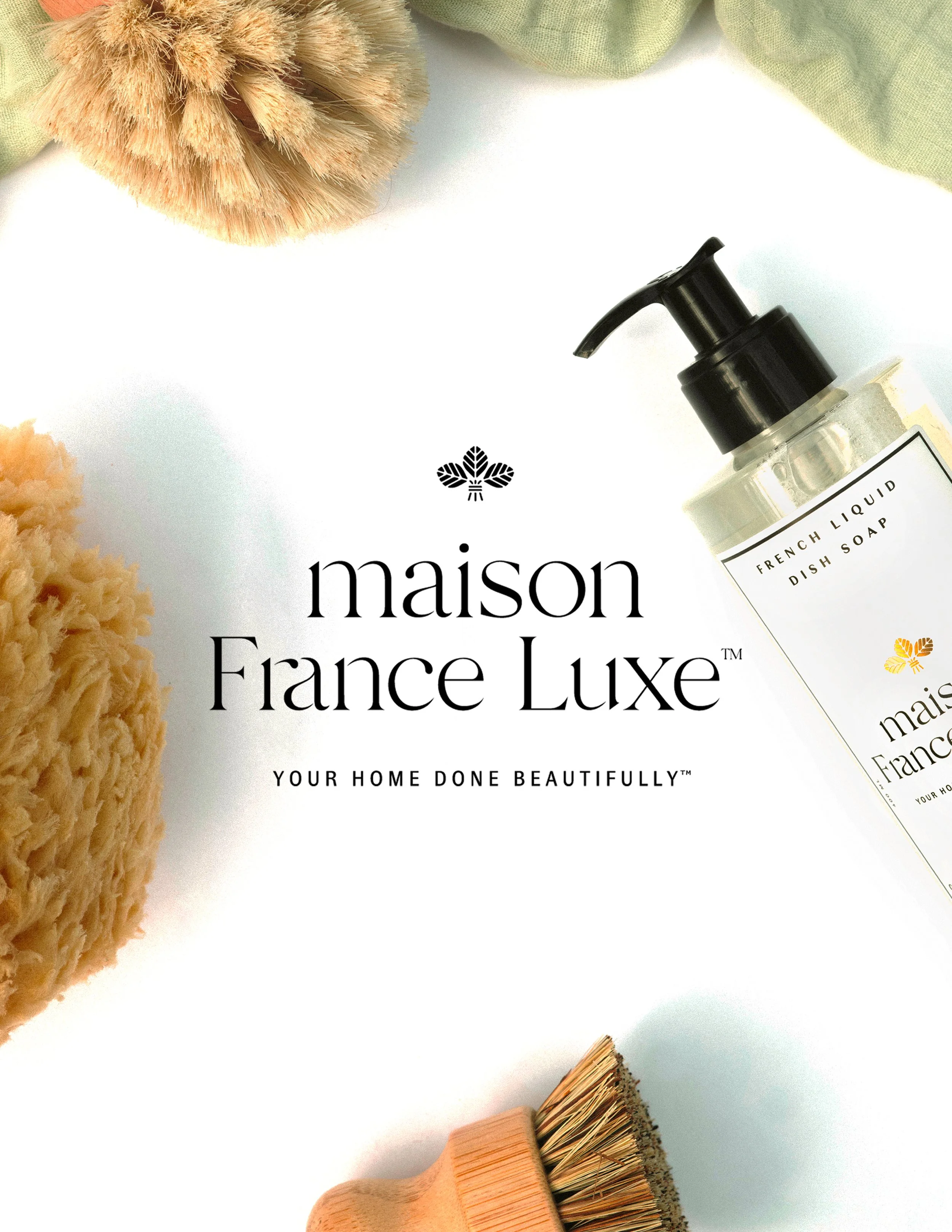 A flat lay of cleaning accessories including a wooden scrub brush, a fluffy beige sponge, and a black pump bottle labeled 'French Liquid Dish Soap.' There are also green cloths and beige textured items around the cleaning supplies, all on a white sur