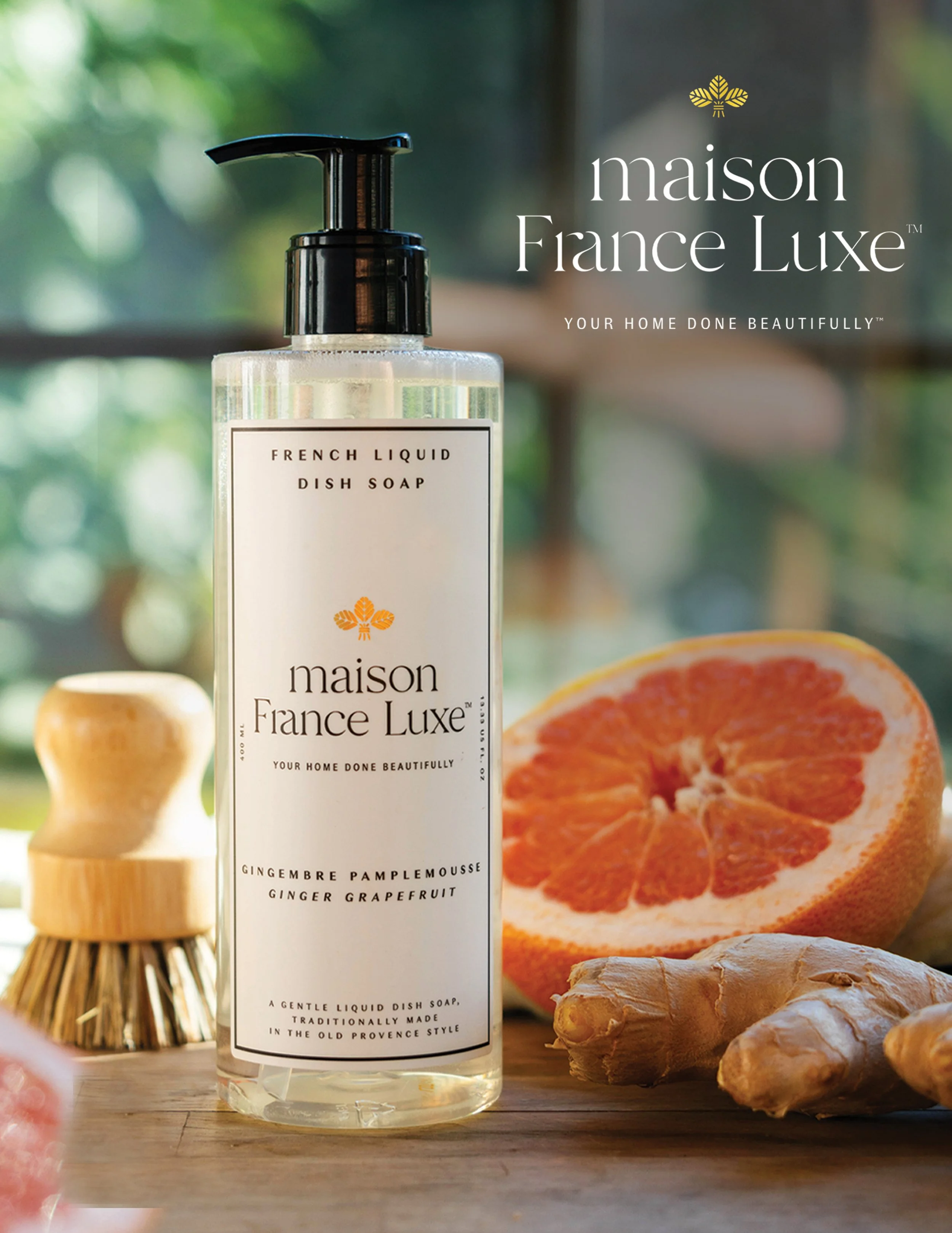 A clear bottle of Maison France Luxe dish soap labeled 'French Liquid Dish Soap Ginger Pamplemousse Ginger Grapefruit' with a black pump, placed on a wooden surface alongside fresh ginger, a halved grapefruit, and a natural-bristled dish brush. The b