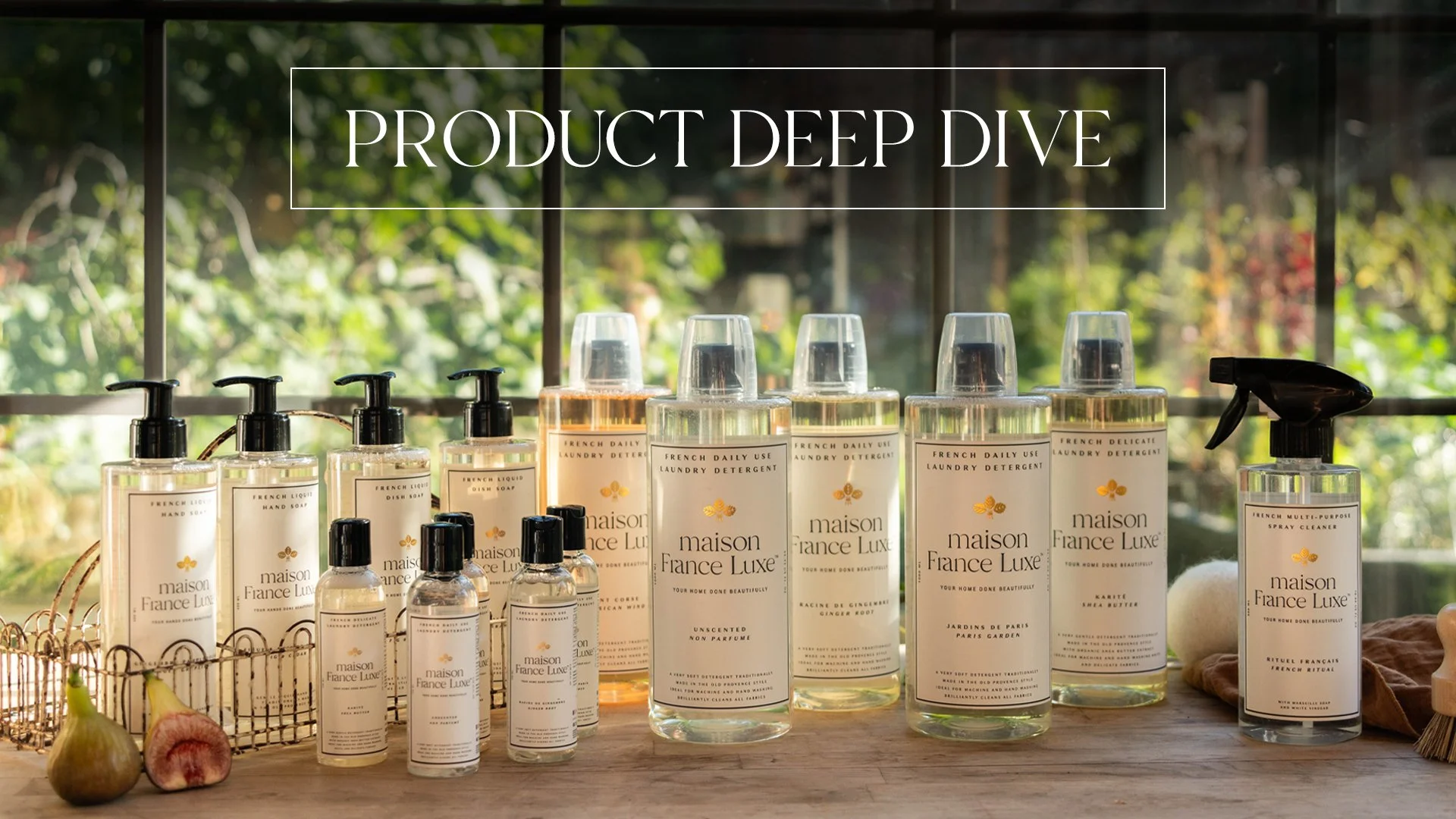 Display of Maison France Luxe laundry and hand soap bottles on a wooden surface, with sliced figs and garlic in the foreground, and a background of green foliage through a window.