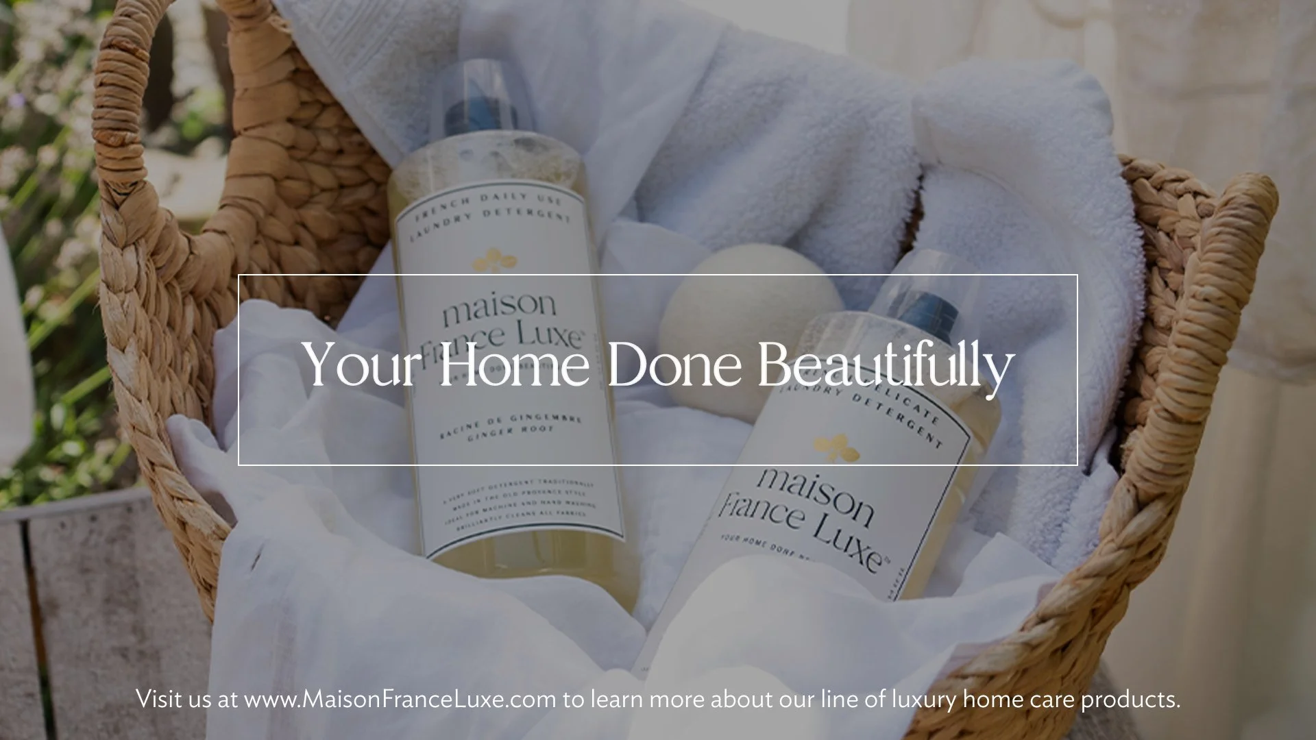 A wicker basket containing two bottles of Maison France Luxe laundry detergent, one is labeled ginger and the other is labeled lavender, along with a rolled white towel and a white bath bomb, all set on white fabric with natural outdoor lighting.