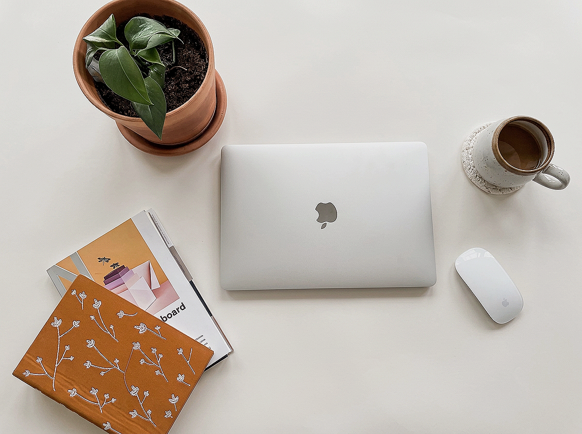 A closed silver MacBook laptop with Apple logo, a white Apple Magic Mouse, a ceramic coffee mug on a coaster, a potted plant with green leaves in a terracotta pot, and two notebooks, one brown with white floral pattern and the other with an illustrated design, all on a white desk.