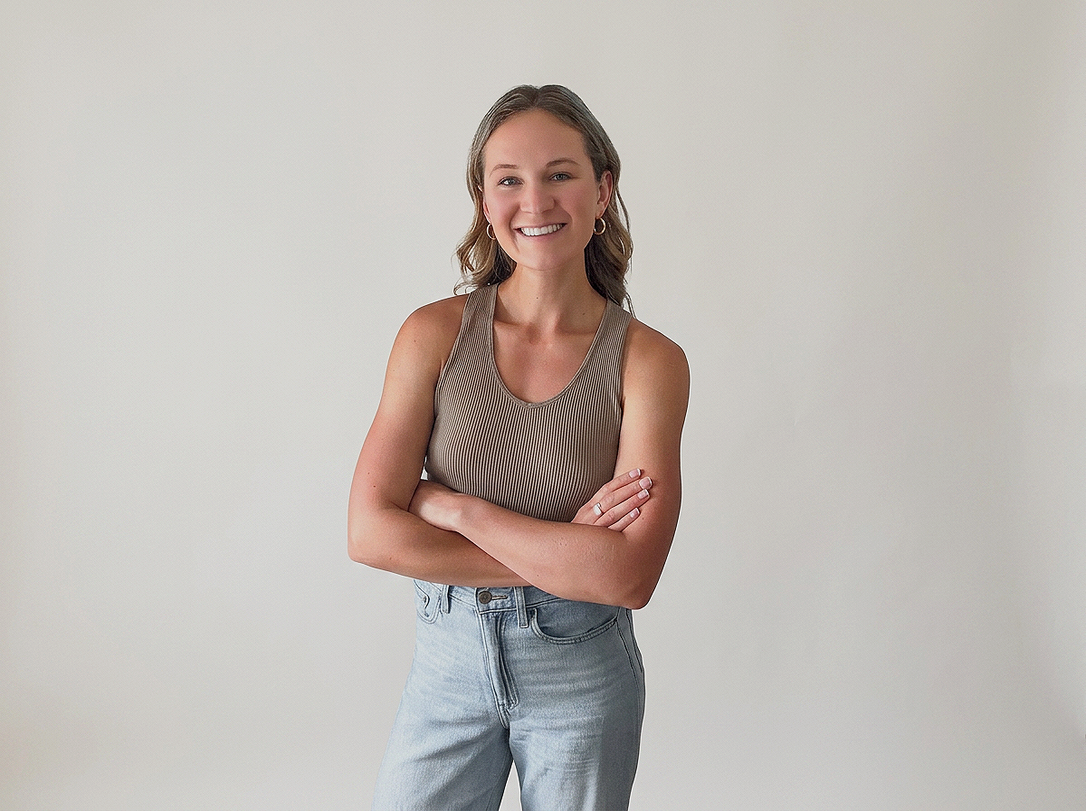 A woman with light brown hair smiling, standing against a plain white wall, wearing a sleeveless ribbed top and high-waisted jeans with arms crossed.