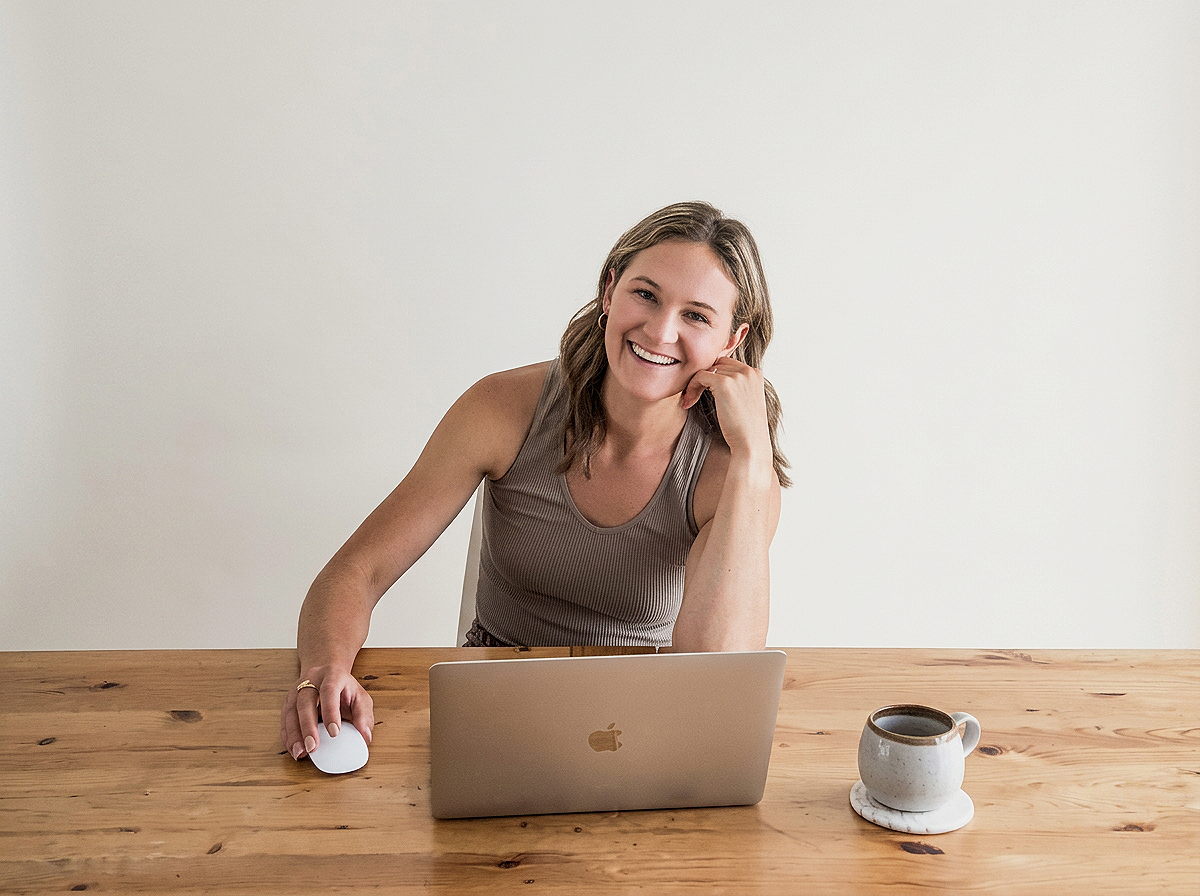 A young woman sitting at a wooden table with a silver MacBook, smiling, with her right hand on a white computer mouse and her left hand resting on her chin, next to a coffee mug.