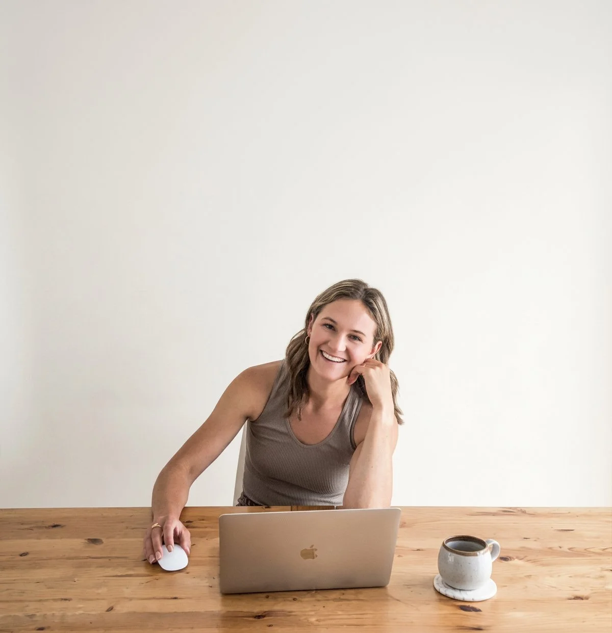 A woman with shoulder-length hair, wearing a sleeveless top, sitting at a wooden desk with a laptop and a cup of coffee, smiling at the camera.