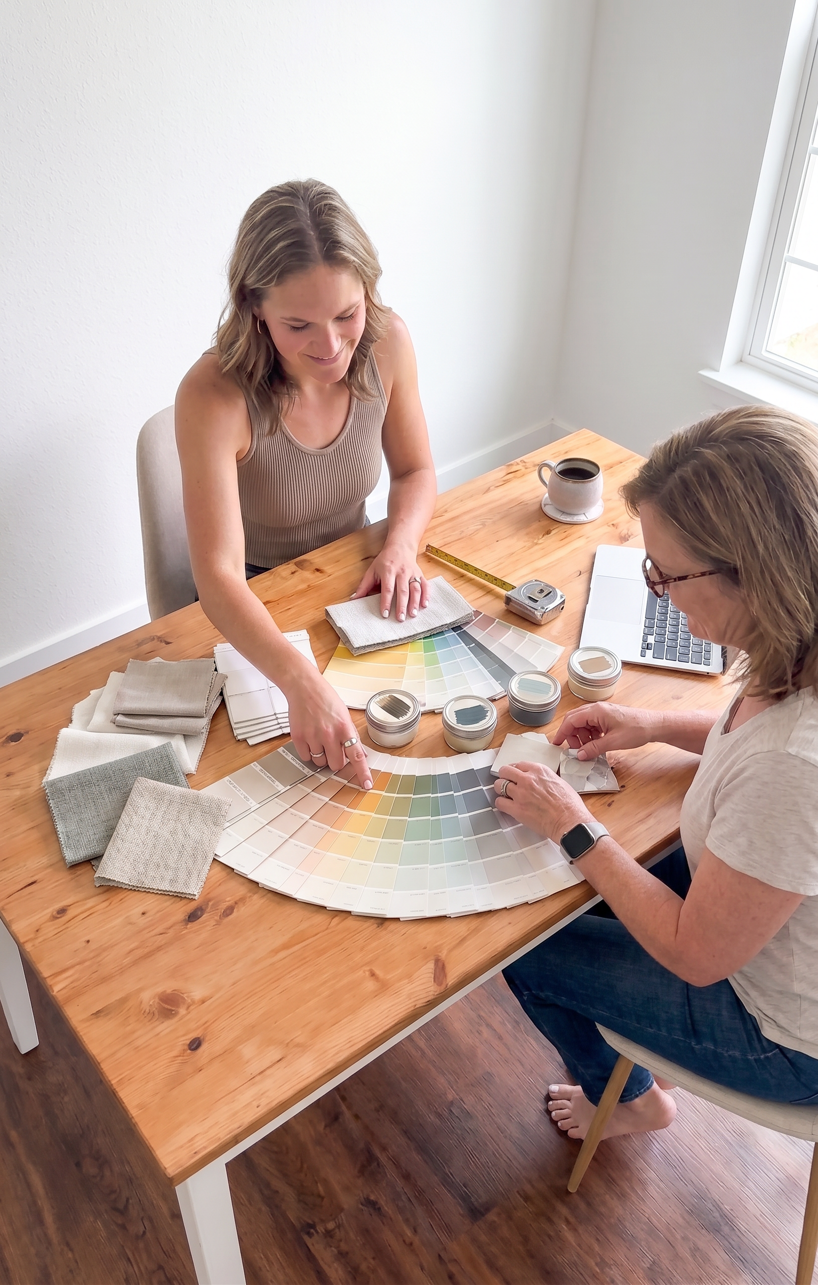 Two women working on a color palette and fabric swatches at a wooden table near a window, with a laptop and coffee mug on the table.