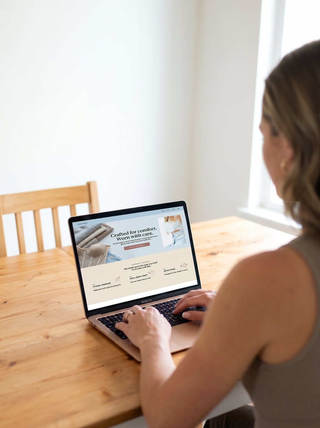 A woman sitting at a wooden table using a MacBook laptop, viewing a website with handmade fabric products.