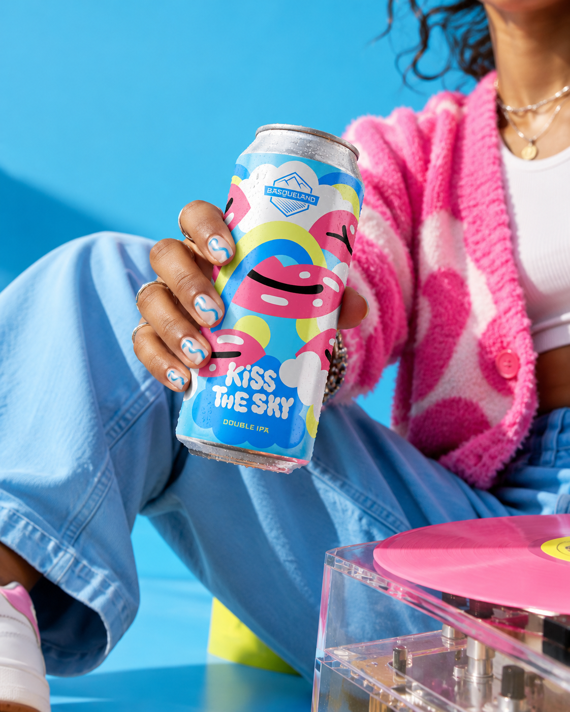 A person wearing light blue jeans, a pink and white striped sweater, and jewelry, holding a colorful can of 'Kiss the Sky' double IPA beer against a bright blue background. There is a pink record and a transparent turntable in front.