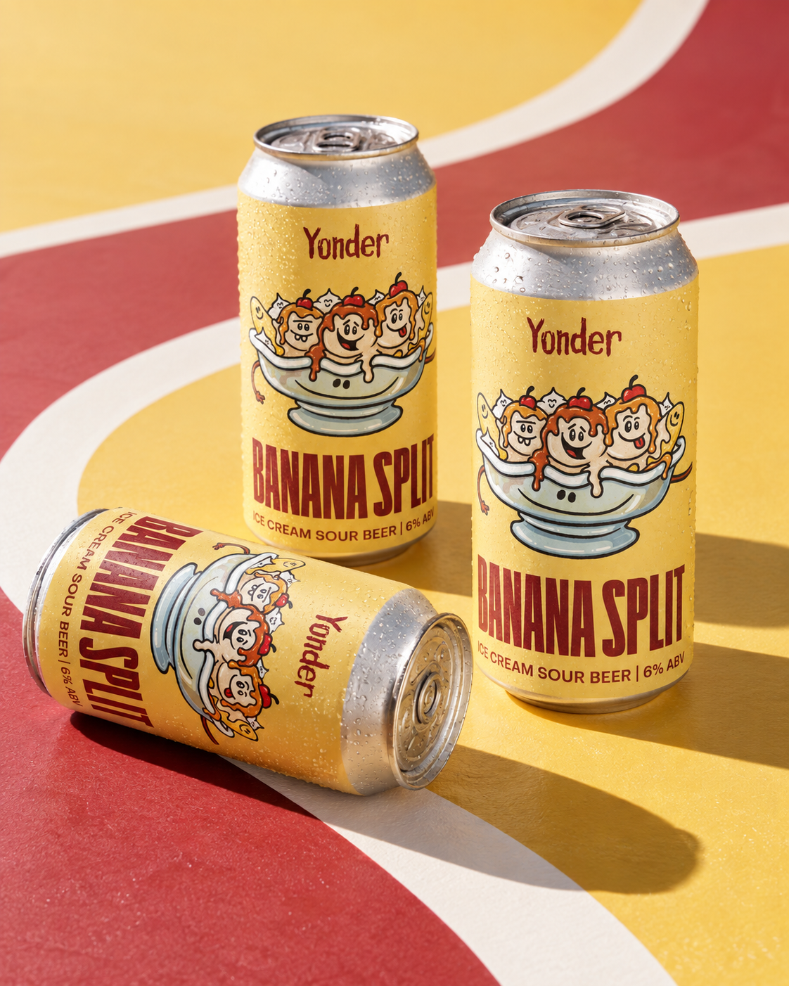 Three cans of Yonder Banana Split ice cream sour beer, with bright yellow labels featuring cartoon characters in a bowl of banana split, condensation on the cans, placed on a colorful surface with red, yellow, and white design.
