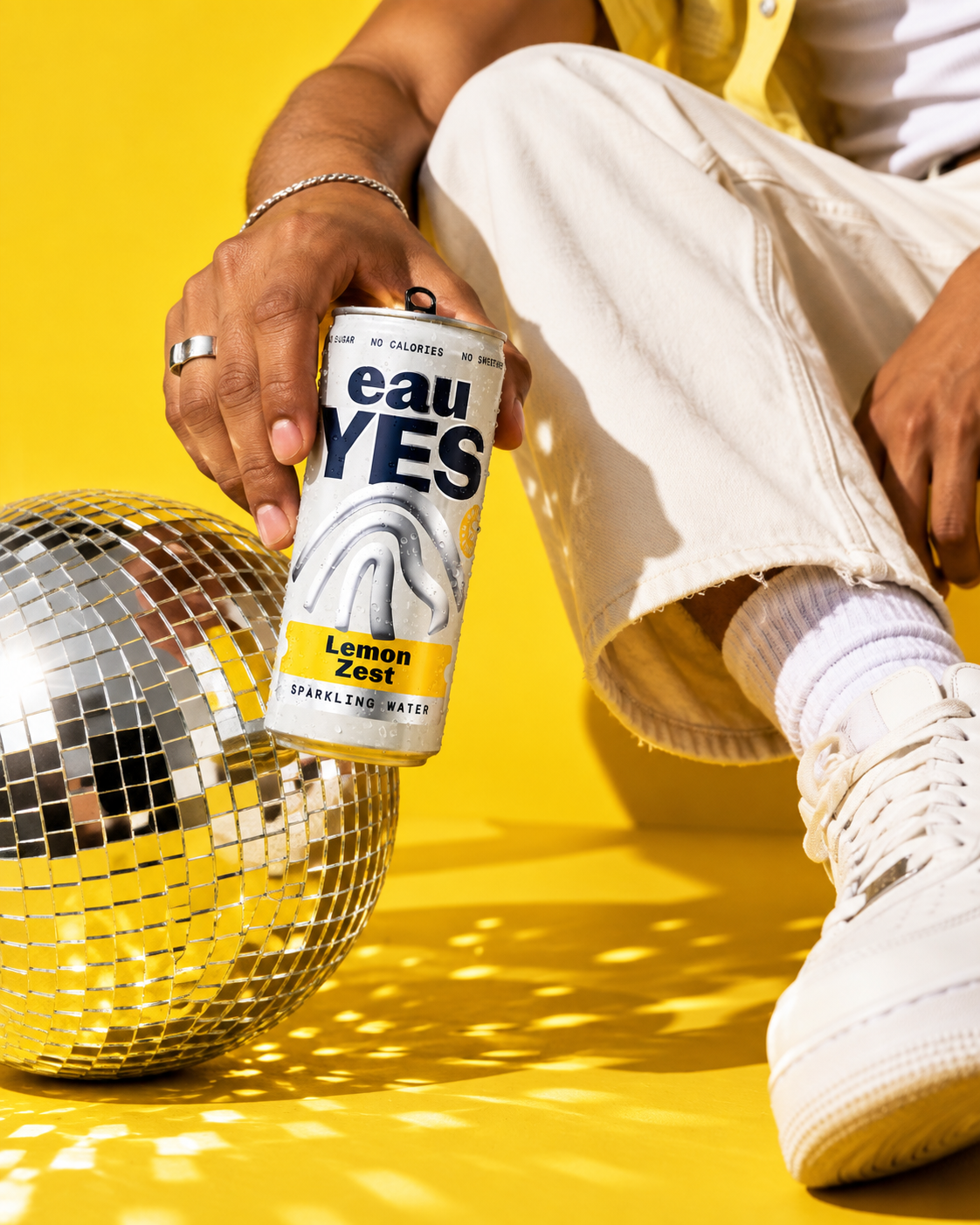 A person wearing beige pants and white sneakers holding a can of Eau Yes Lemon Zest sparkling water, kneeling beside a small disco ball on a bright yellow background.