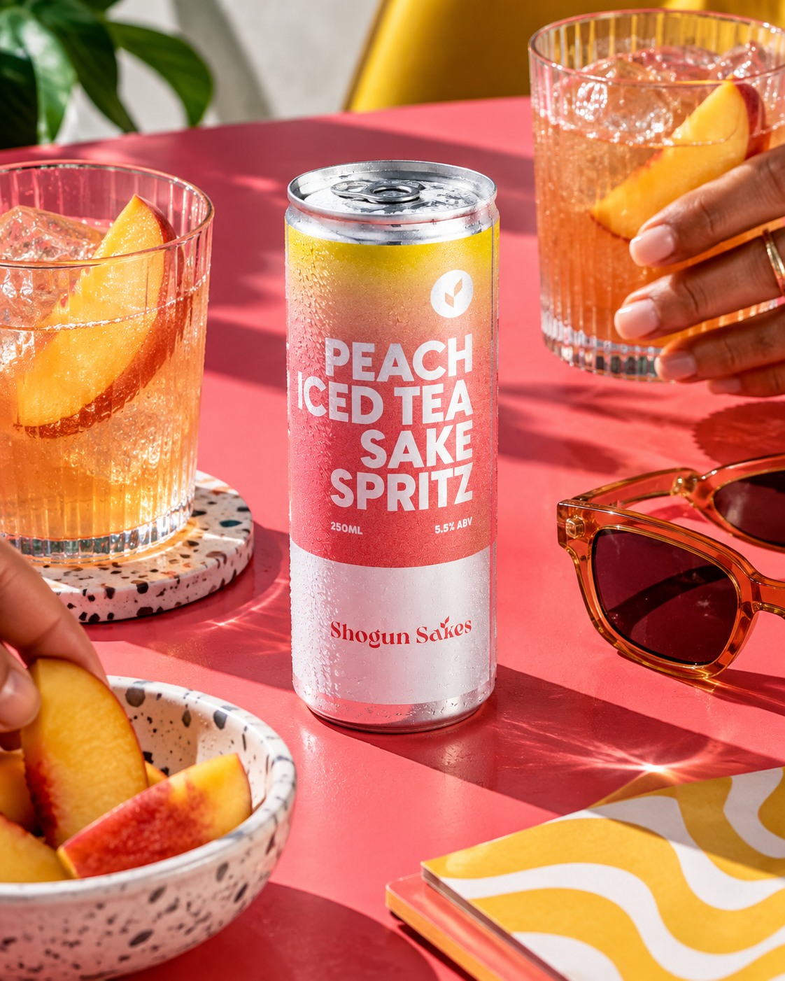 A can of Peach Iced Tea Sake Spritz on a pink table, with two glasses filled with the beverage and peach slices, a bowl of peach slices, sunglasses, and a patterned notebook.