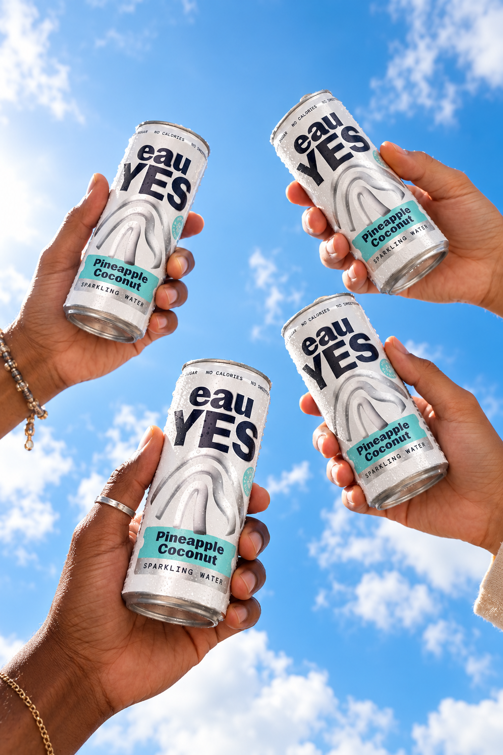 Four hands holding cans of Eau Yes Pineapple Coconut sparkling water are raised against a blue sky with scattered clouds.
