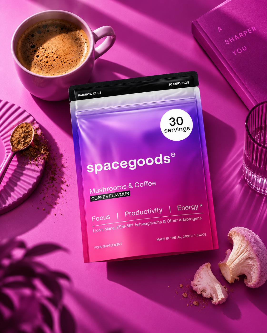 A package of spacegoods Mushrooms & Coffee supplement on a pink surface surrounded by a pink coffee mug, a pink dish with a spoon, pieces of mushrooms, a glass of water, a purple book, and a clear glass, with shadows and pink lighting.