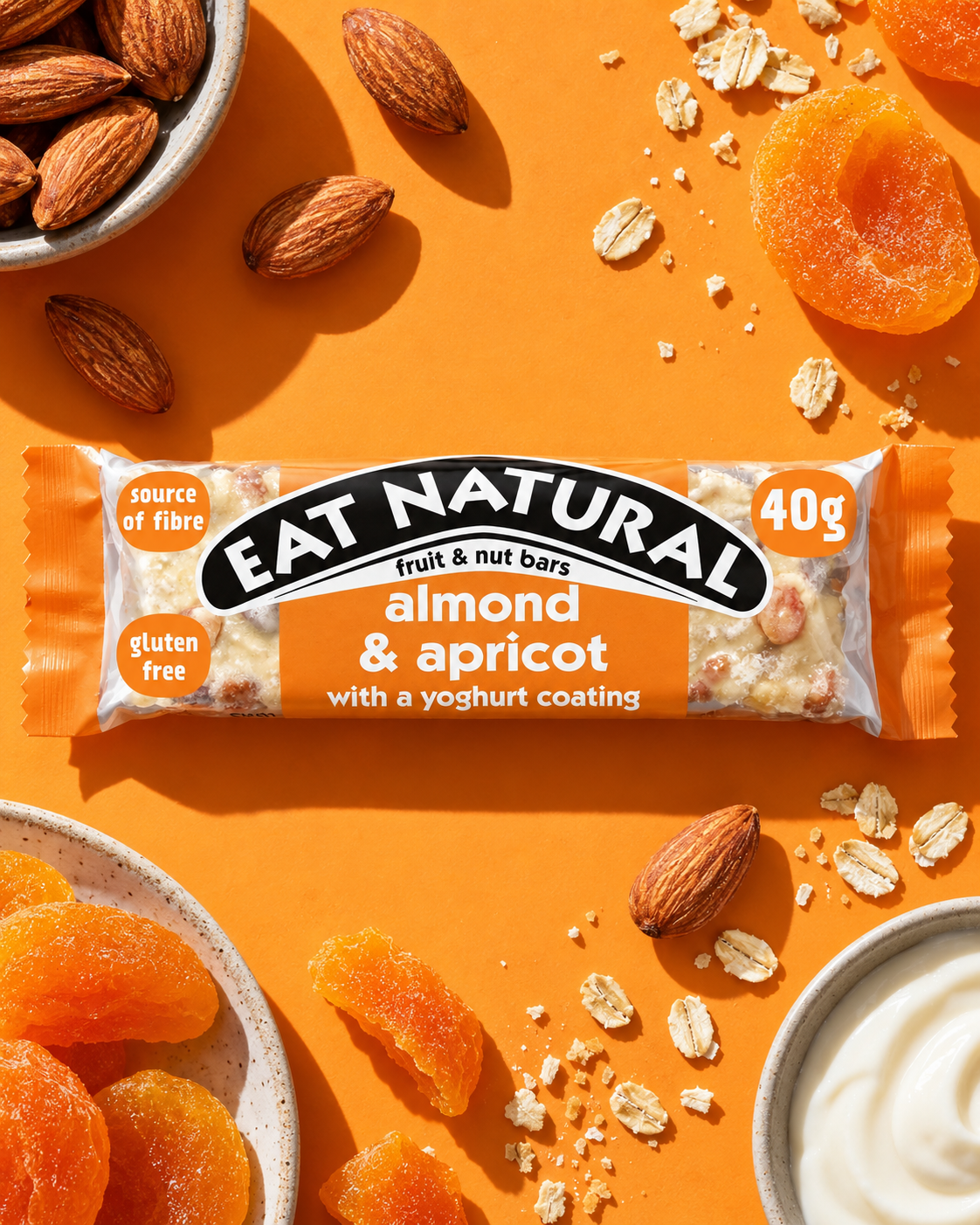 A fruit and nut bar labeled 'Eat Natural' with almond and apricot flavor, placed on an orange surface. Surrounding the bar are almonds, dried apricots, oats, and a bowl of yogurt.