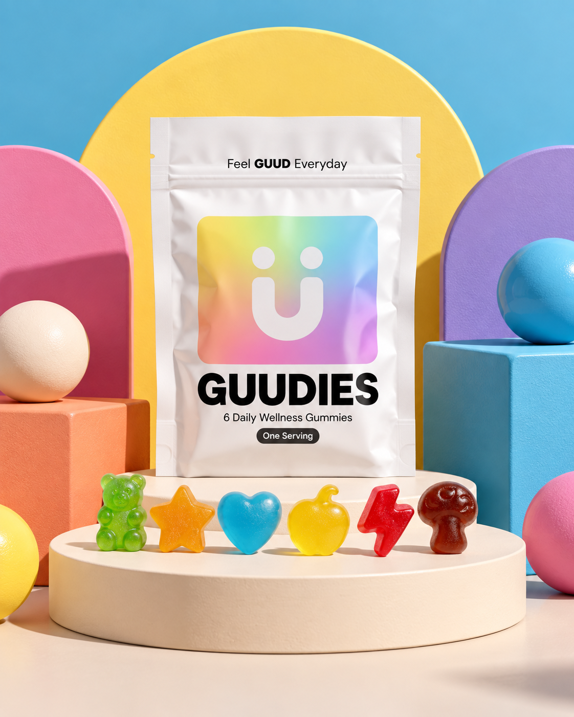 Brightly colored wellness gummies and candies arranged on a white circular platform, with a package labeled 'GUUDIES' in the center, surrounded by pastel-colored geometric shapes and spheres.