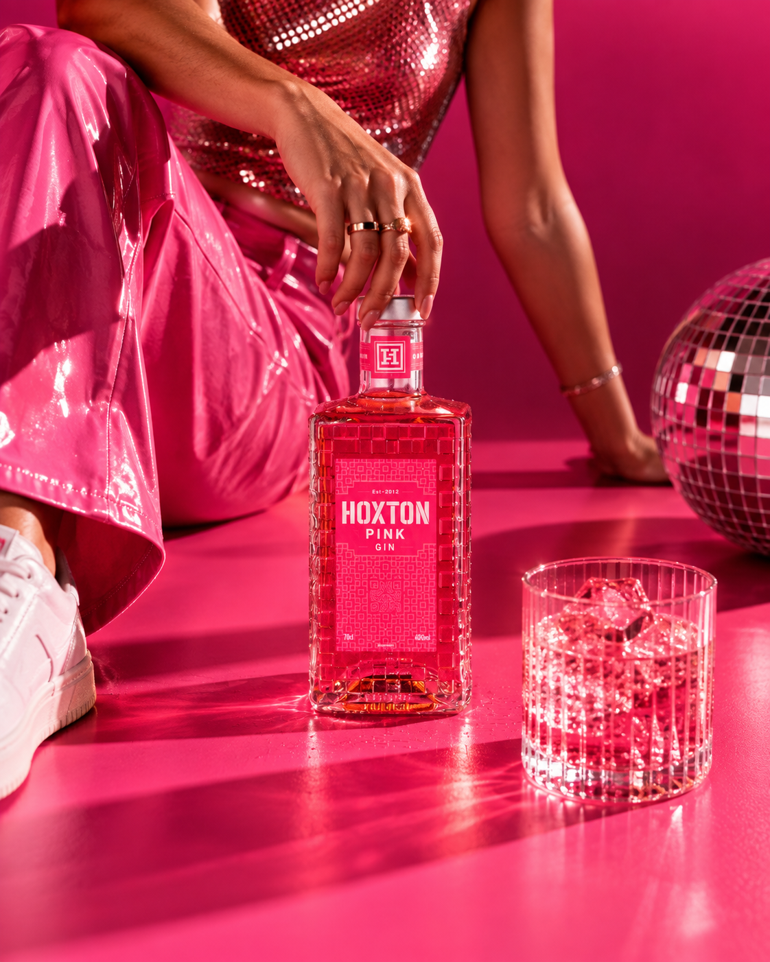 A person dressed in pink shiny clothing is reaching for a pink Gin bottle labeled 'Hoxton Pink Gin' on a pink surface. There is a crystal bowl with ice and a disco ball in the background.