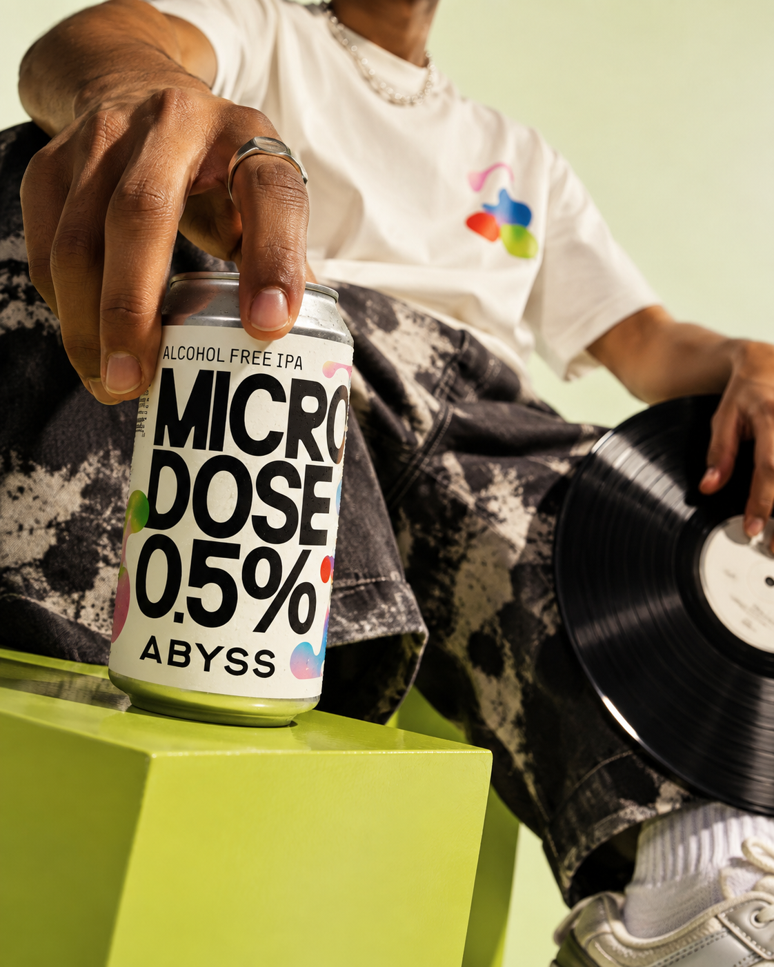 A person holding a can of Microdose Alcohol Free IPA with a vinyl record in their other hand, sitting on a bright green surface.