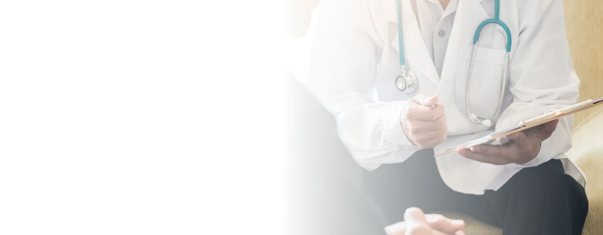 A healthcare professional in a white coat with a stethoscope around their neck is writing on a clipboard.  A patient is sitting in front of them.