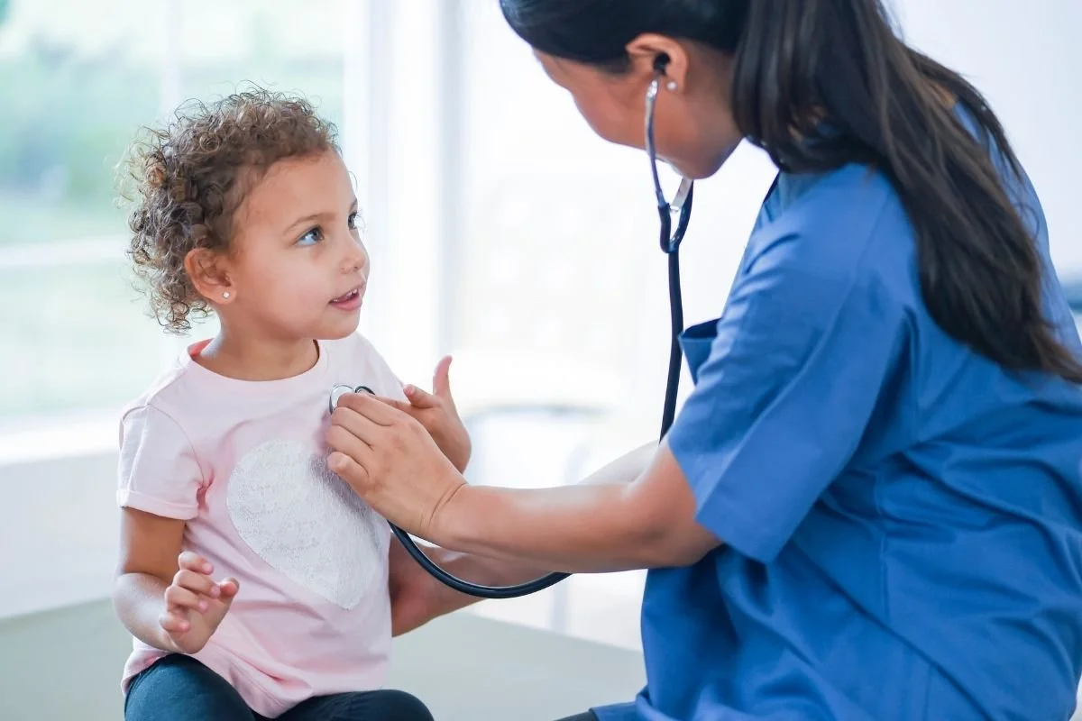A young girl talking to a healthcare professional, a nurse or doctor, who is using a stethoscope to listen to her chest.