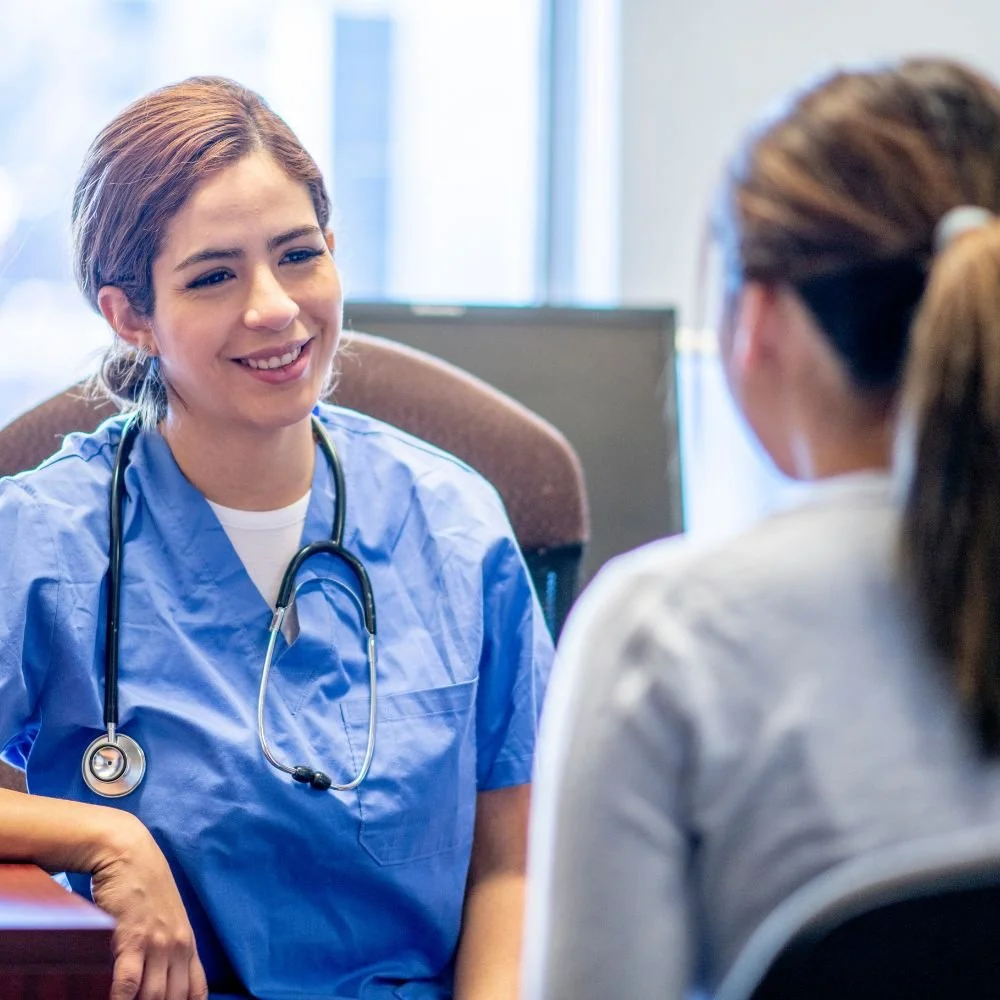 A young female healthcare professional wearing blue scrubs and a stethoscope, smiling and talking to a patient in a medical office.