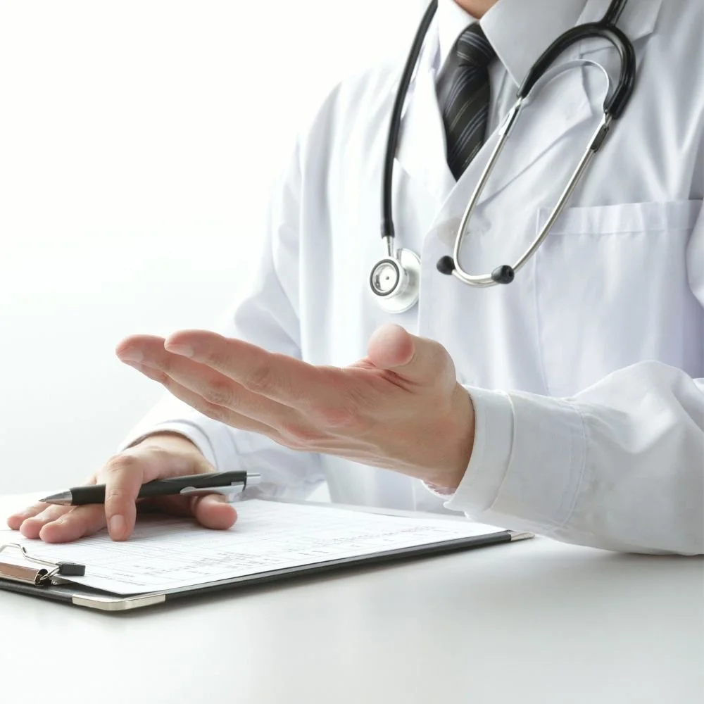A doctor in a white coat with a stethoscope around his neck, holding a pen and gesturing with his hand, sitting at a desk.