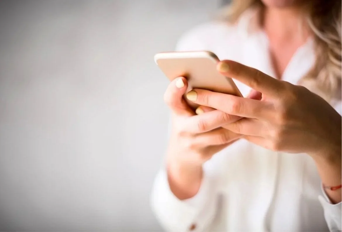A woman using a smartphone with painted nails, partially visible in a white shirt, against a plain background.