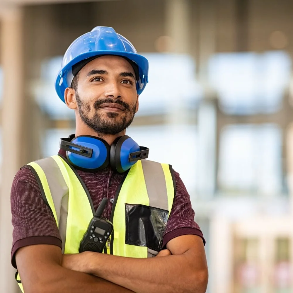 A construction worker wearing a blue safety helmet, a high-visibility vest, and ear protection, standing in an indoor construction site with blurred background.