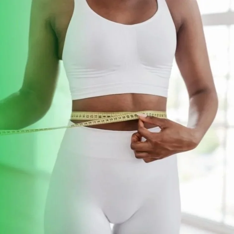 Person measuring their waist with a yellow measuring tape, wearing a white sports bra and white leggings, against a brightly lit background.