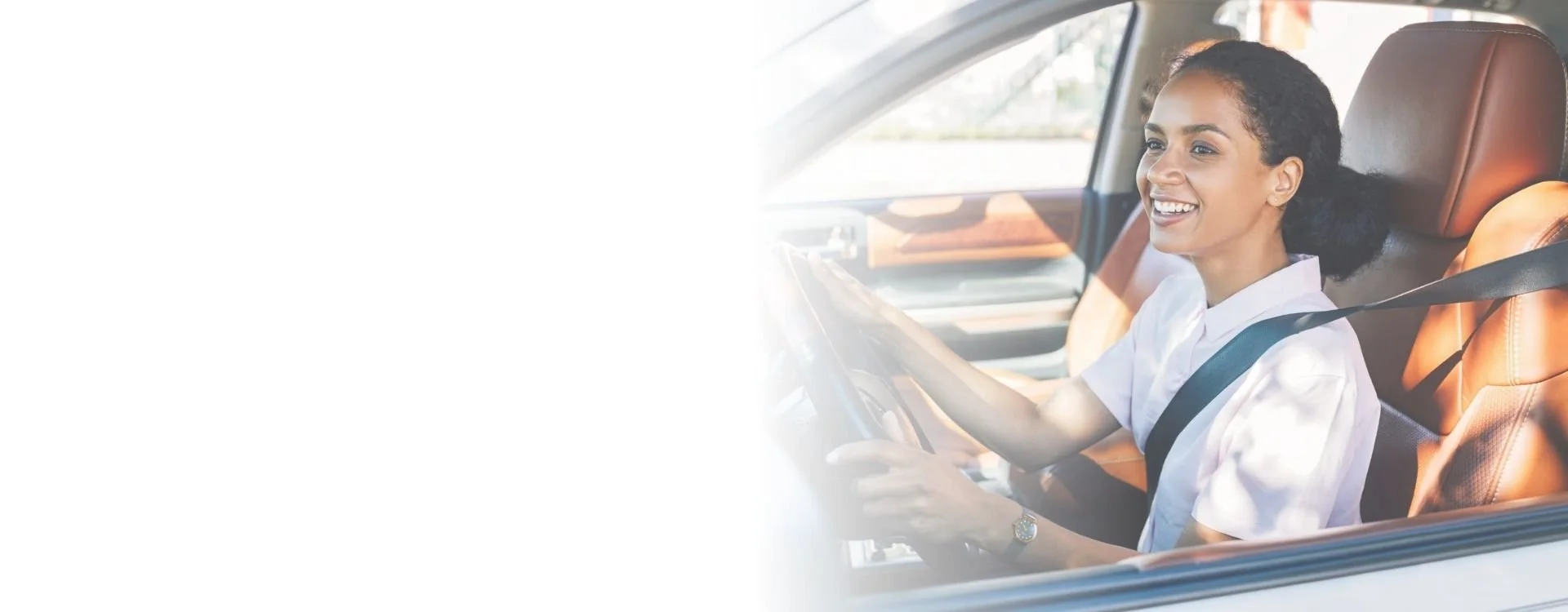 Smiling woman driving a car with a seatbelt on, seated in the passenger seat, sunlight coming through the windshield.
