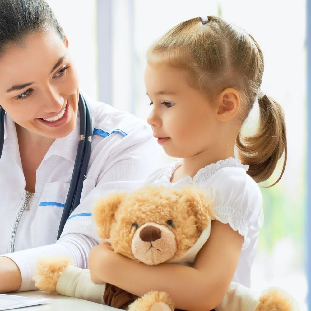 A young girl sitting with a teddy bear, talking to a female doctor or nurse who is smiling at her.