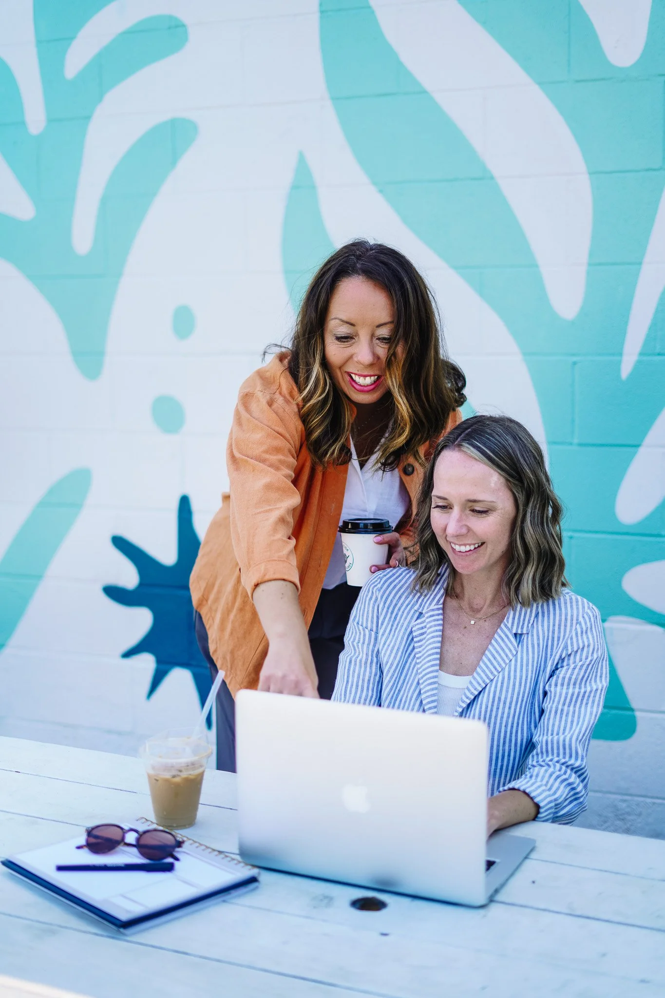 Two women, one standing and one sitting at a table outdoors, working on a laptop with a blue and teal abstract mural in the background. The woman standing has brown hair and is holding a coffee cup. The woman sitting has shoulder-length brown hair and is wearing a light blue striped blazer. Items on the table include sunglasses, a notebook, and a cold coffee beverage.