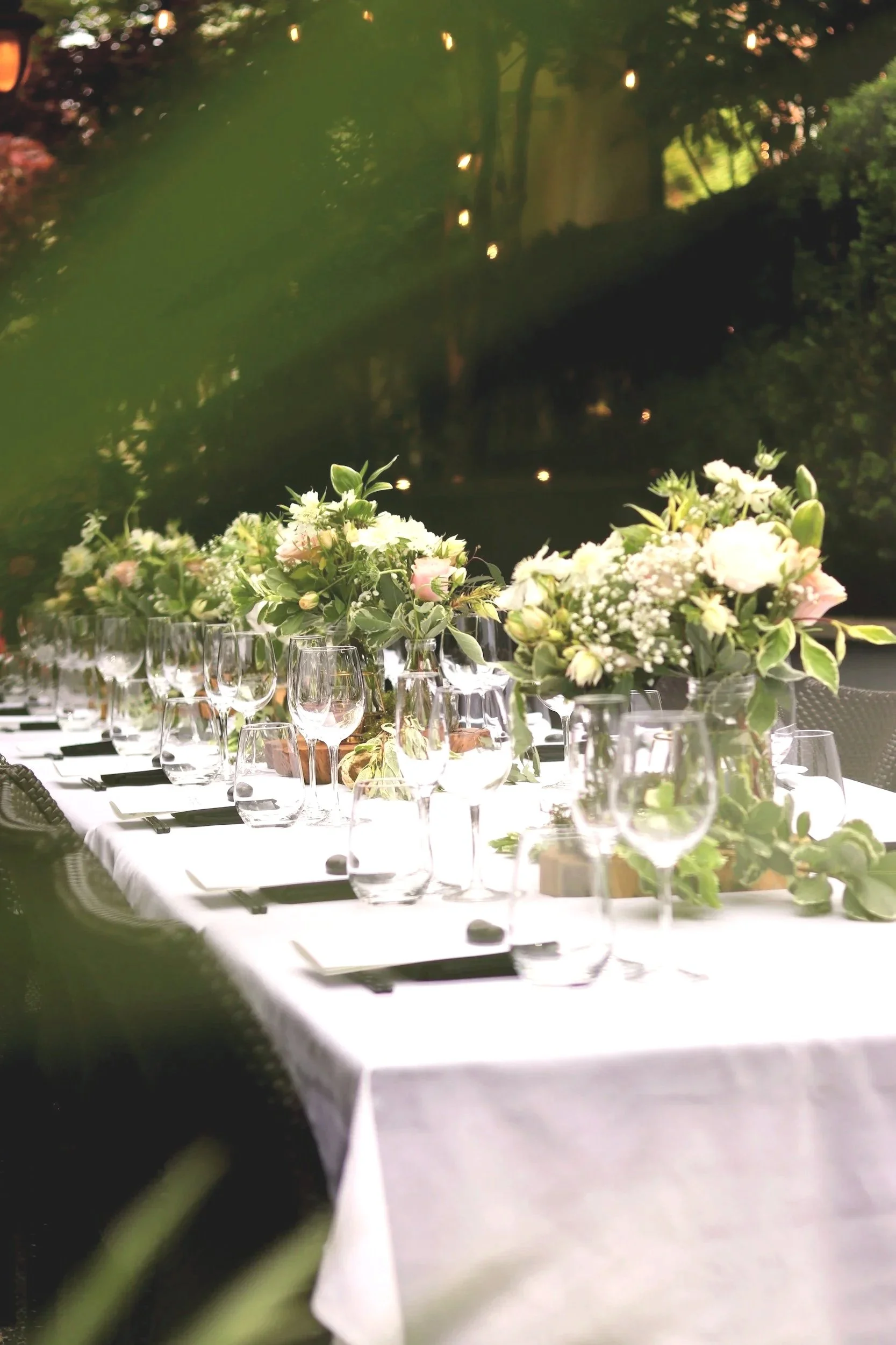 A long banquet table with a white tablecloth decorated with floral centerpieces, multiple wine glasses, and place settings, set outdoors at dusk with trees and lights in the background.