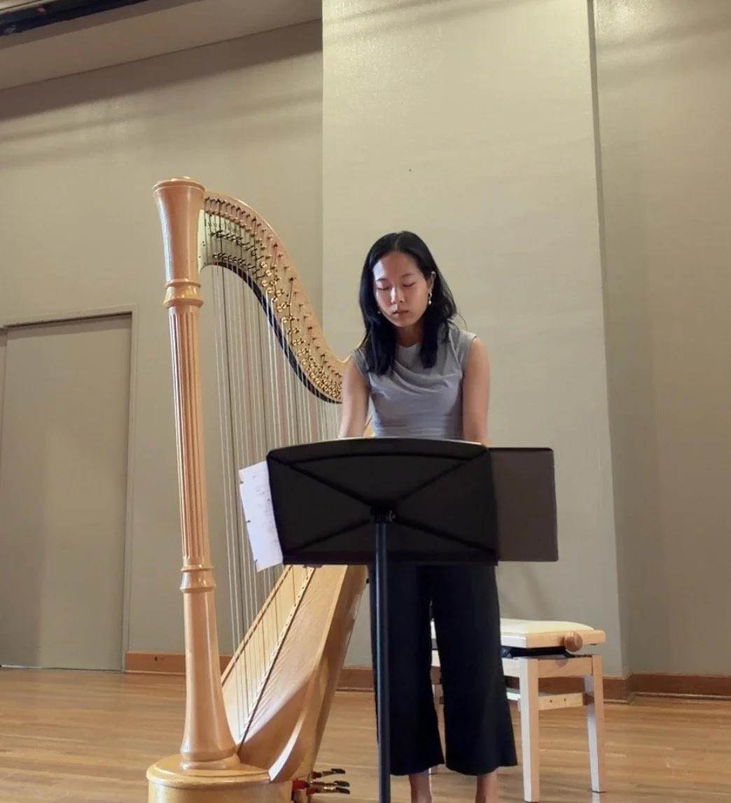 A young woman playing a harp in a room with wooden floor, music stand, and chair.