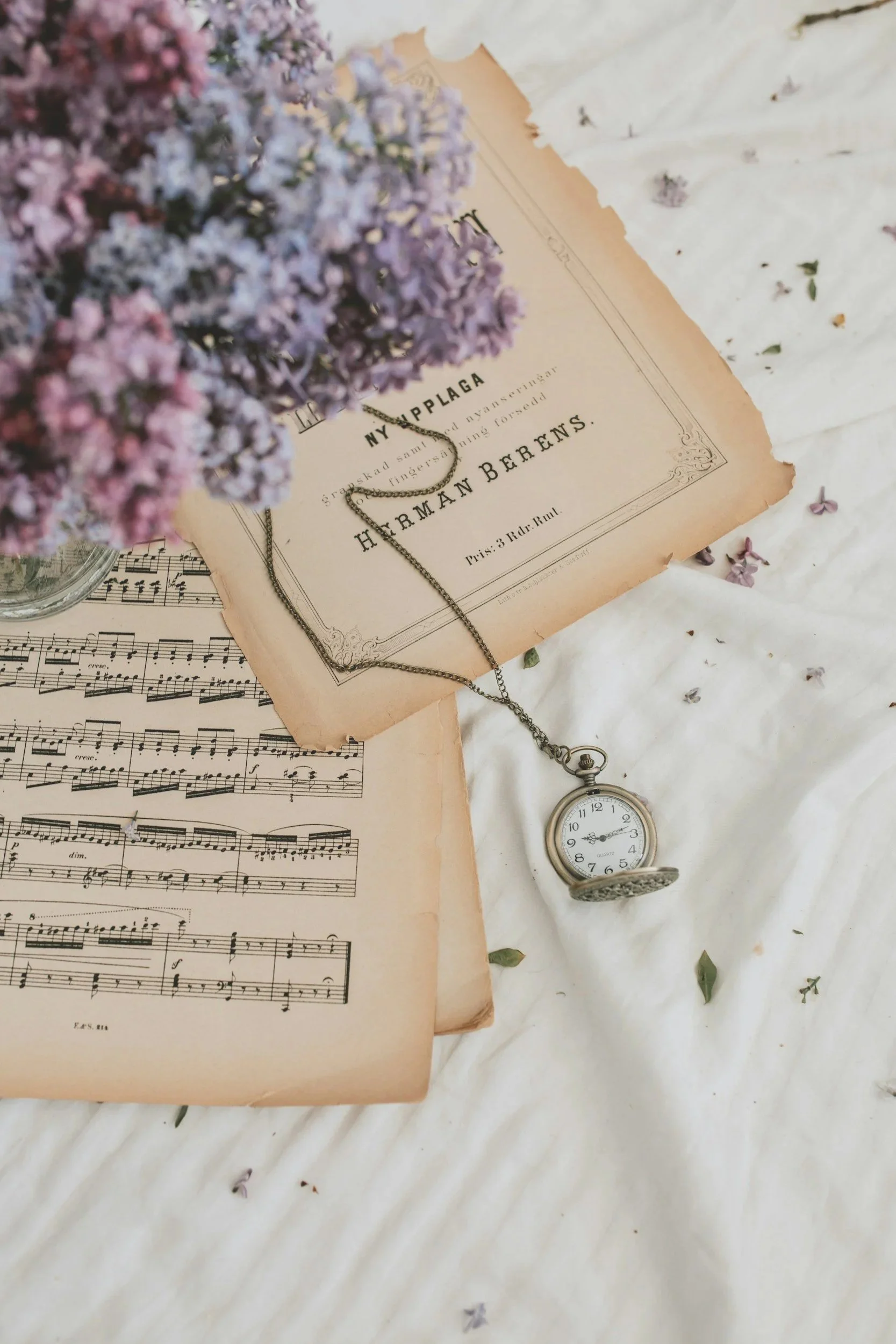 Close-up of a vintage pocket watch on a white quilted fabric, with sheet music and a torn sheet of paper, purple flowers, and a necklace.