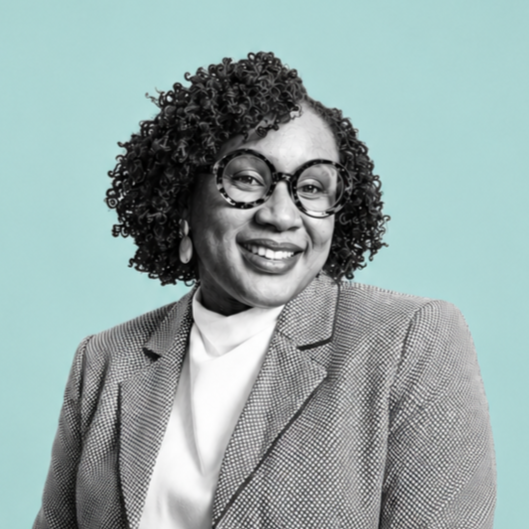 Portrait of a woman with curly hair wearing glasses, a blazer, and a white top, smiling against a light turquoise background.