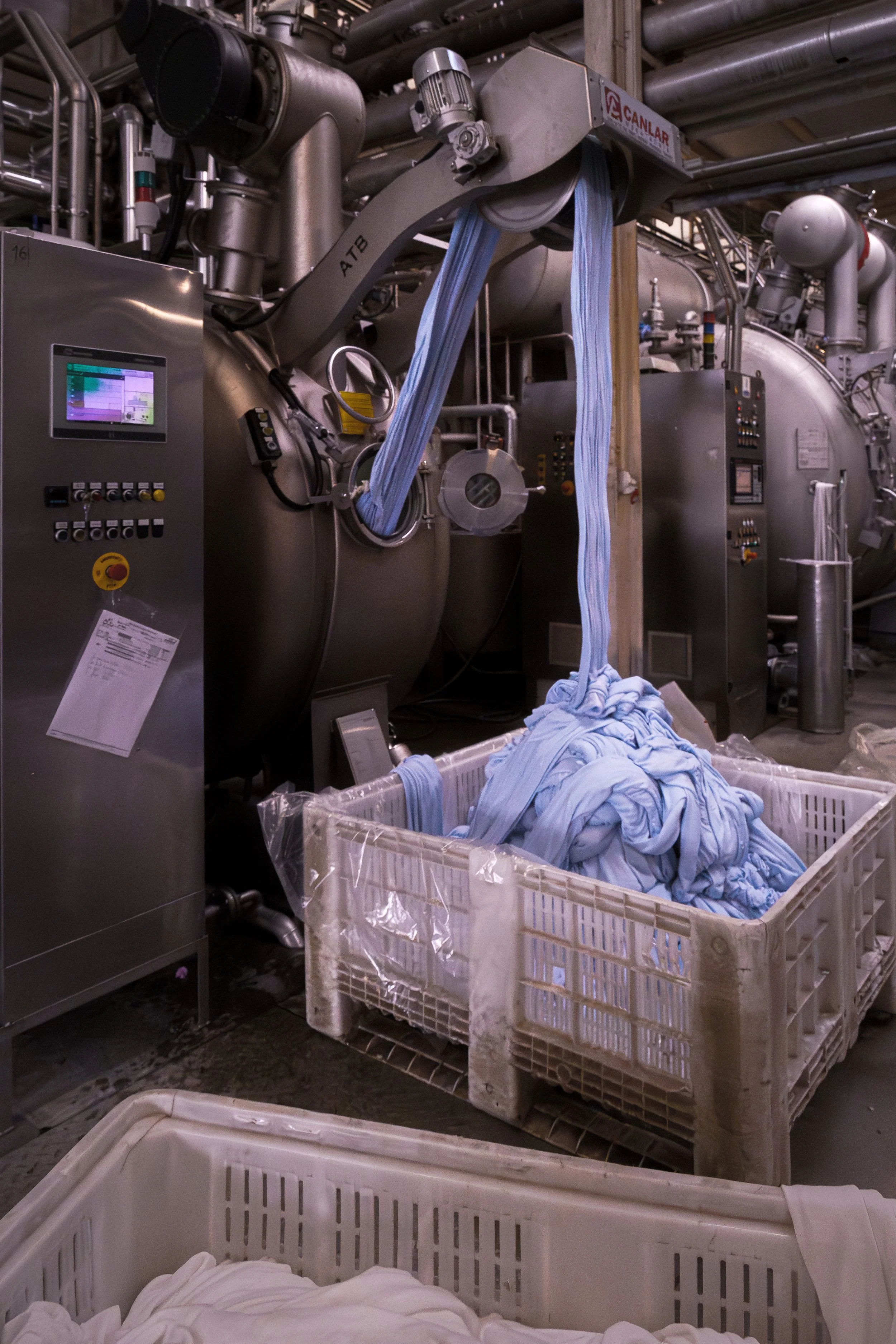 Industrial laundry facility with a large machine processing light blue textiles, with bound fabric hanging from the machine and piles of folded laundry in a plastic basket nearby.