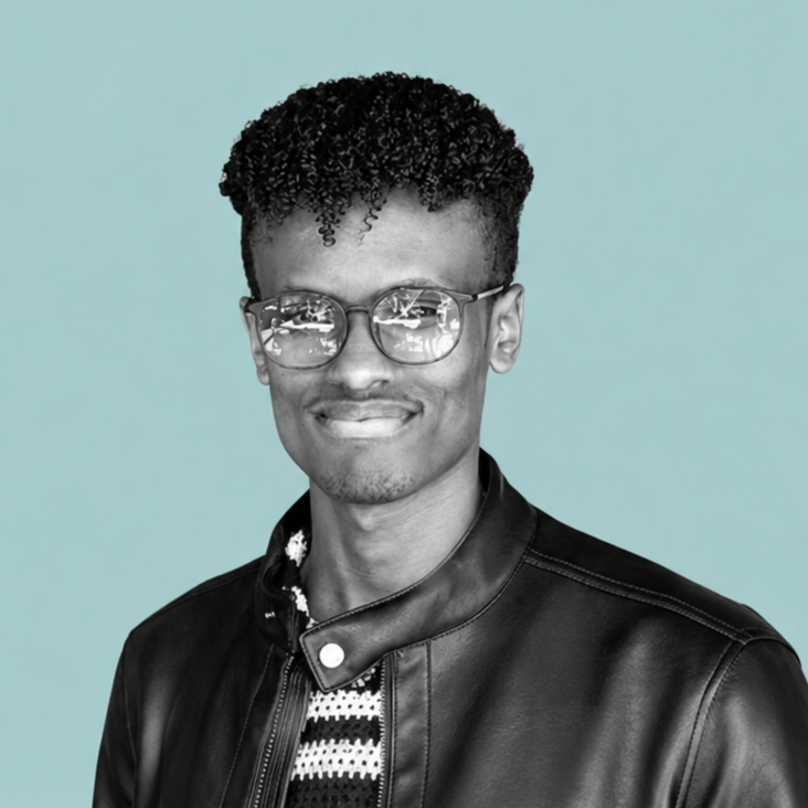 Portrait of a young man with curly hair, glasses, and a leather jacket, smiling against a light blue background.