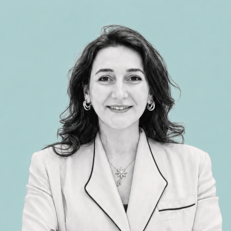 Black and white portrait of a woman with wavy hair, wearing earrings, a necklace, and a light-colored blazer with dark piping, smiling at the camera against a plain background.