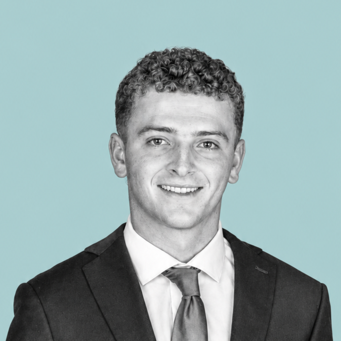 A young man with curly hair, wearing a suit and tie, smiling against a light blue background.